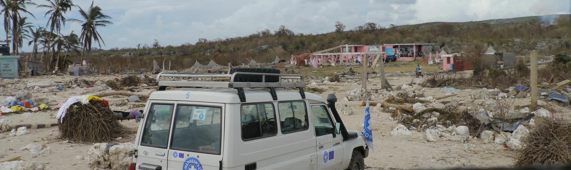 Een wagen met het logo van Dokters van de Wereld staat geparkeerd temidden van een verwoest landschap, huizen in puin, ontbrekende daken, sommige helemaal met de grond gelijk gemaakt.