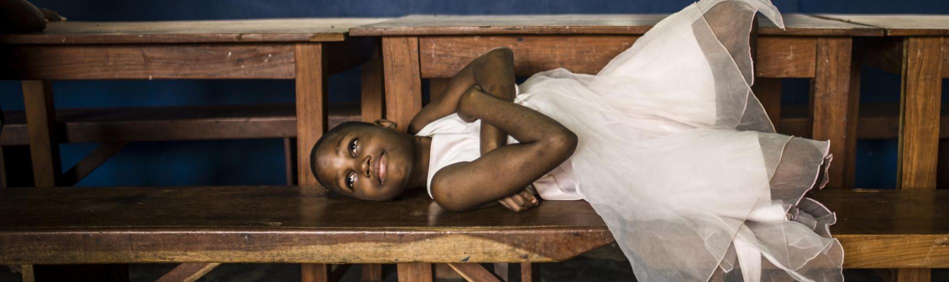 Een meisje uit Benin ligt uitgestrekt op een houten schoolbank. Ze draagt een wit zomerjurkje, heeft de armen gekruist over haar borst en tilt haar hoofd op. Haar blik lijkt licht geamuseerd.