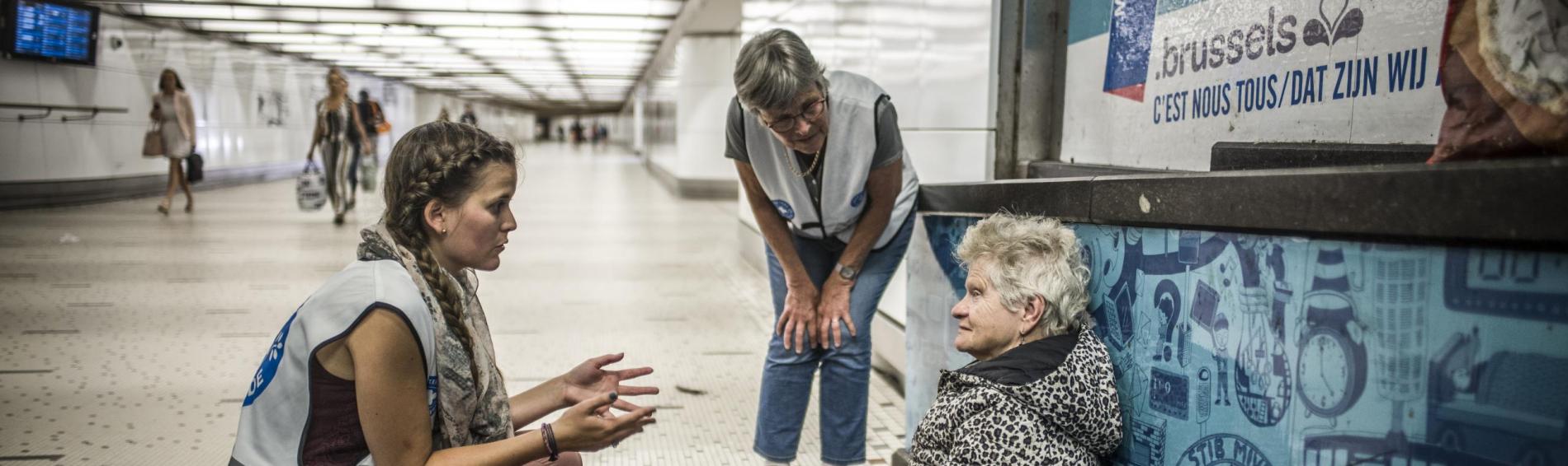 Twee medewerksters van Dokters van de Wereld zijn aan het werk in een metrohal. Ze zijn in gesprek met een oudere vrouw die op de grond zit bovenop een krant. Ze heeft kort wit haar en draagt een dikke, lange winterjas boven een roze jurk en wandelschoenen.