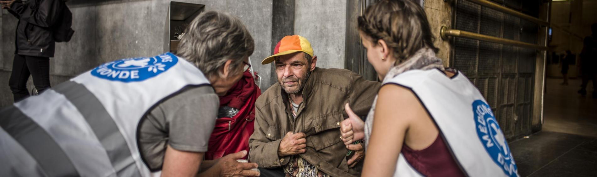 Voor het Noordstation in Brussel zitten twee medewerksters van Dokters van de Wereld gehurkt op de grond naast een dakloze man. Ze leggen hem uit wat onze organisatie voor hem kan doen. 