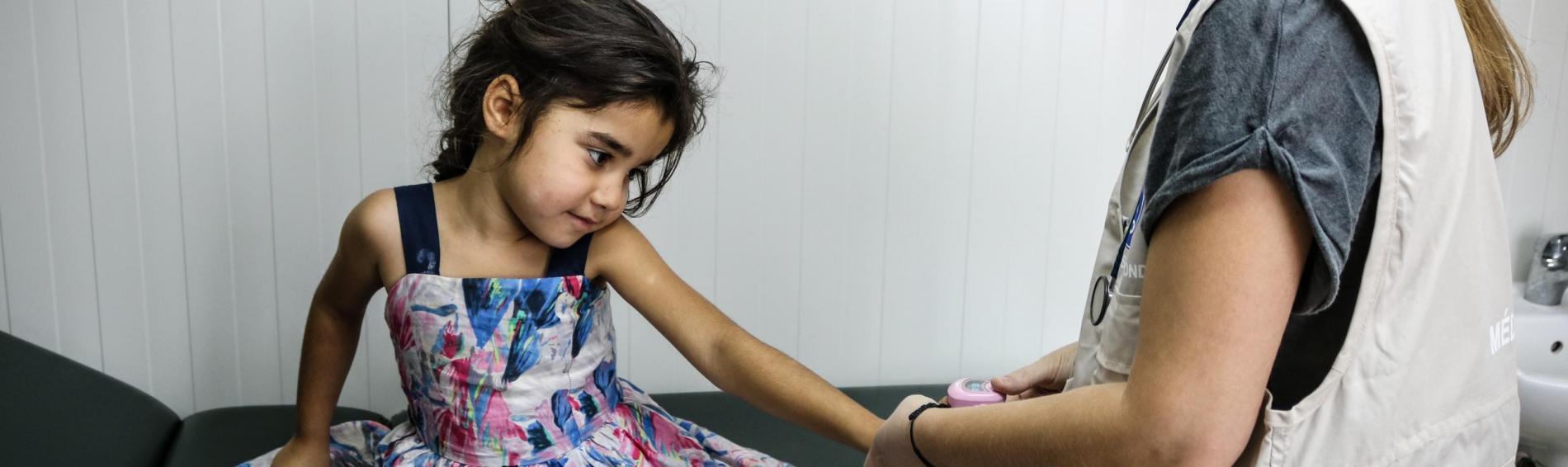 Een Syrisch meisje in kleurrijke jurk met bloemetjes zit op een dokterstafel in een Grieks vluchtelingenkamp. De Griekse kinderdokter van Dokters van de Wereld meet haar bloeddruk.