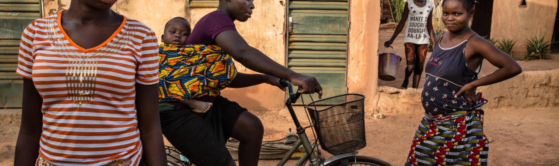 Drie jonge Burkinese vrouwen in een straat in Ouagadougou. Eén van hen zit op een fiets met haar baby in een doek om haar rug gebonden.