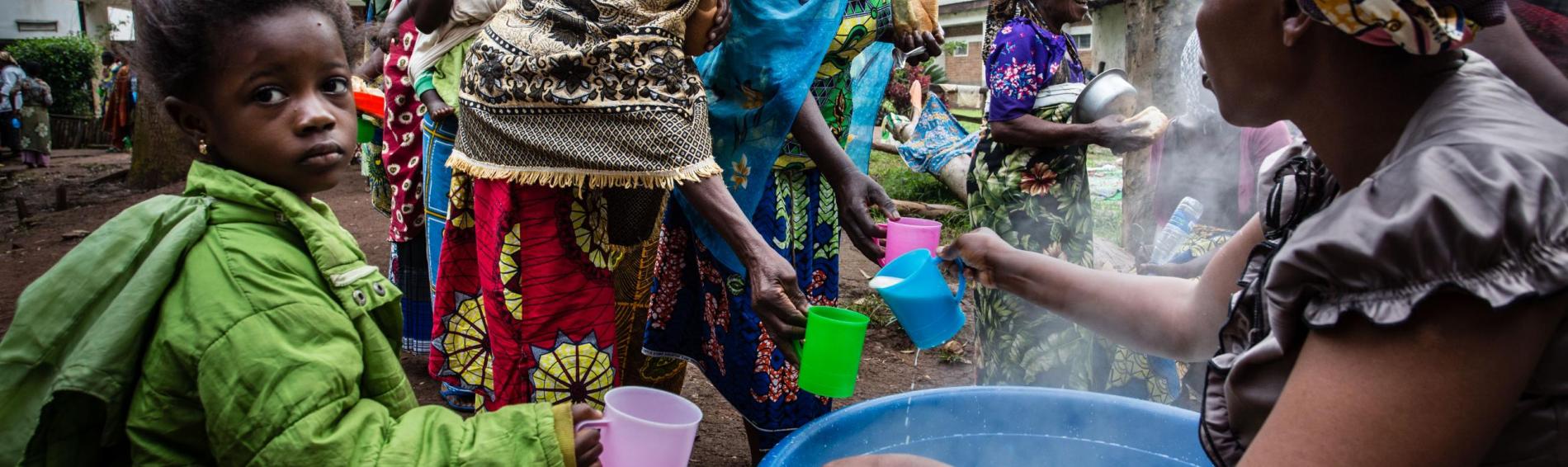 Congolese vrouwen en kinderen in kleurrijke jurken en hoofddoeken vullen hun bekers met drinkwaterwater in een plastieken teil. Eén van hen heeft de teil op haar schoot en kijkt toe dat de verdeling eerlijk verloopt. 