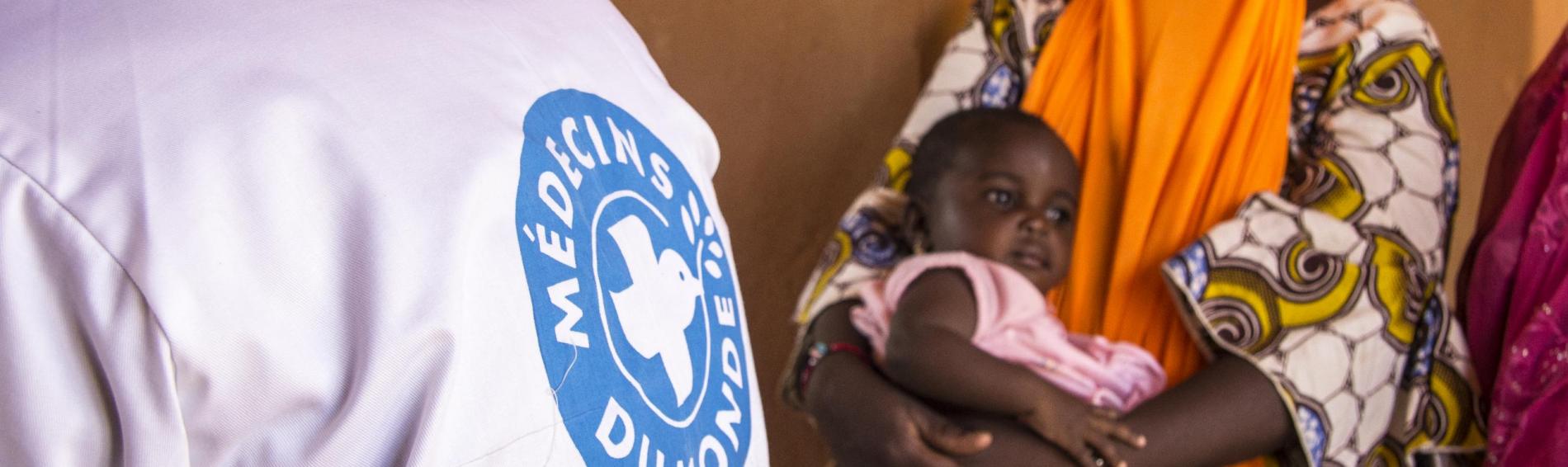 Een Malinese baby in de armen van haar moeder. Op de voorgrond één van onze artsen met een jas waarop het logo staat.