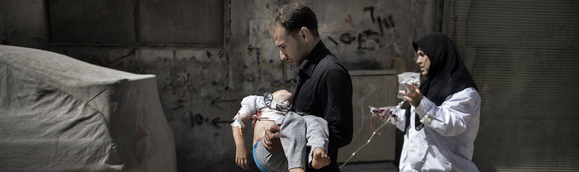 Een Syrische vader draagt zijn kind in de armen door straten die in puin liggen. Achter hen loopt een vrouw met hoofddoek hen op de voet met een bakster die aan het kind is bevestigd.