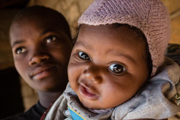 Een jonge Congolese mama houdt haar baby in haar armen. Het kind kijkt recht in de lens, mond samengevouwen alsof het een zoen wil geven en één wenkbouw omhoog getrokken.