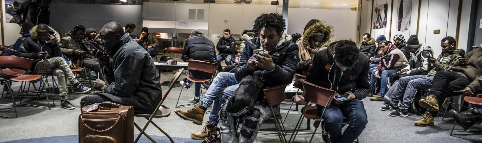 Een groep migranten in de wachtkamer van de humanitaire hub. Door hun smartphones scrollen verlicht het wachten, vooral dankzij de Refugee Aid app dat met steun van Dokters van de Wereld die aan mensen onderweg in België informatie verstrekt die hun het leven iets makkelijker maakt.