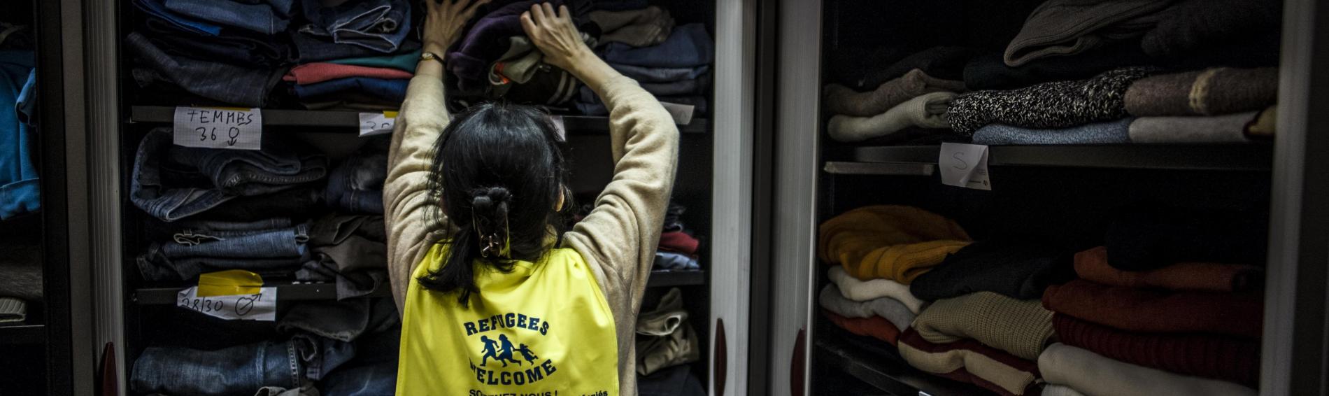 Een vrijwilligster met een geel jasje waarop 'refugees welcome' gemarkeerd staat, houdt zich bezig met het sorteren van te doneren kledij in het magazijn van de humanitaire hub. De kast puilt uit van de kleurrijke wintertruien, warme broeken en jassen die per maat zijn uitgestald.