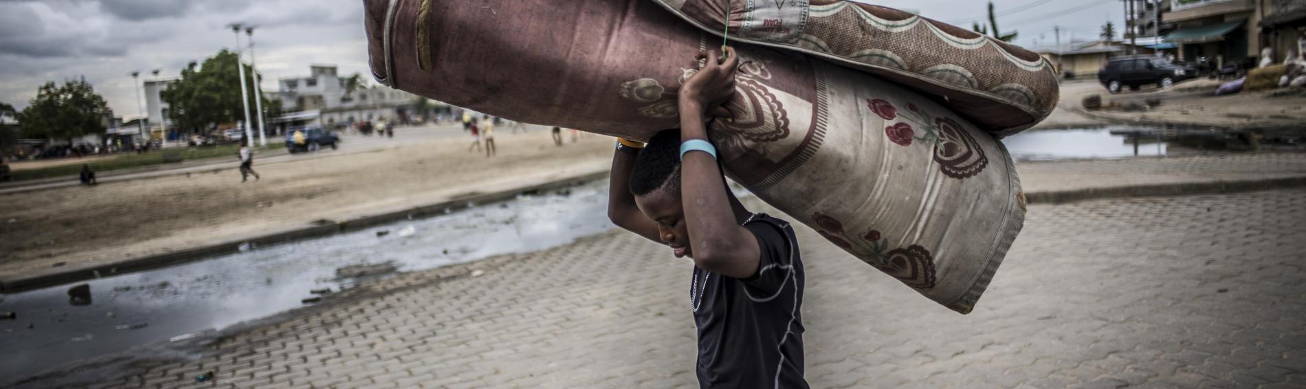 Een tienerjongen op straat in Benin. Het is bewolkt, hij draagt een vochtig, dichtgeplooid matras boven zijn hoofd. Op de achtergrond spelende kinderen in een verlaten buitenwijk.