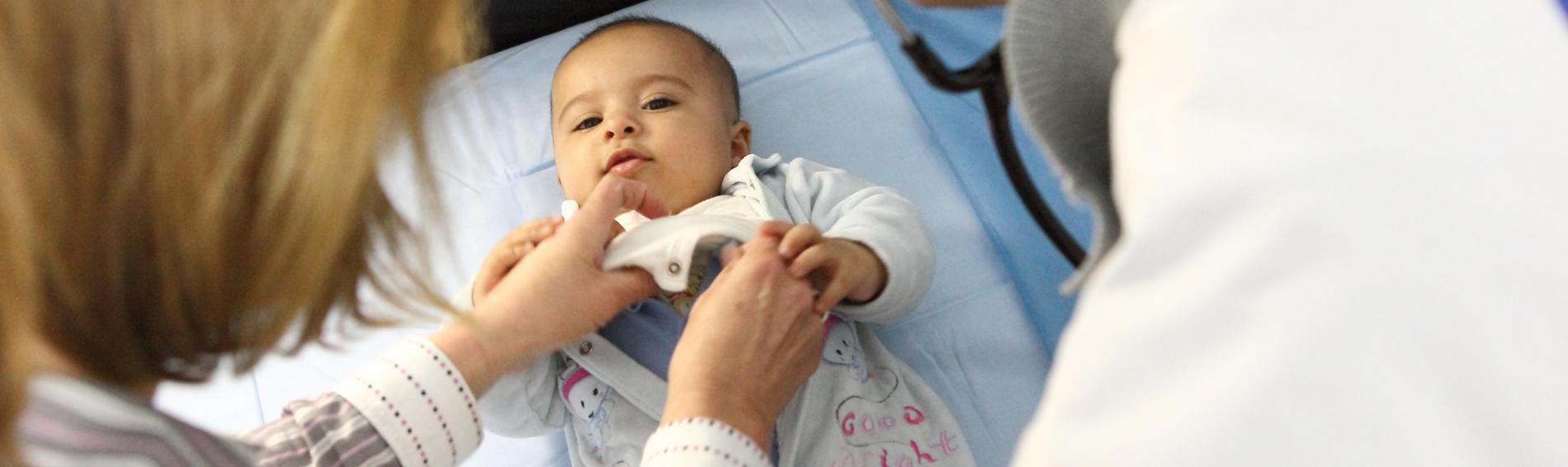 Een dokter met een witte jas waarop ons logo staat onderzoekt met zijn stethoscoop een baby die recht in de lens kijkt terwijl de mama haar kind tracht te kalmeren.
