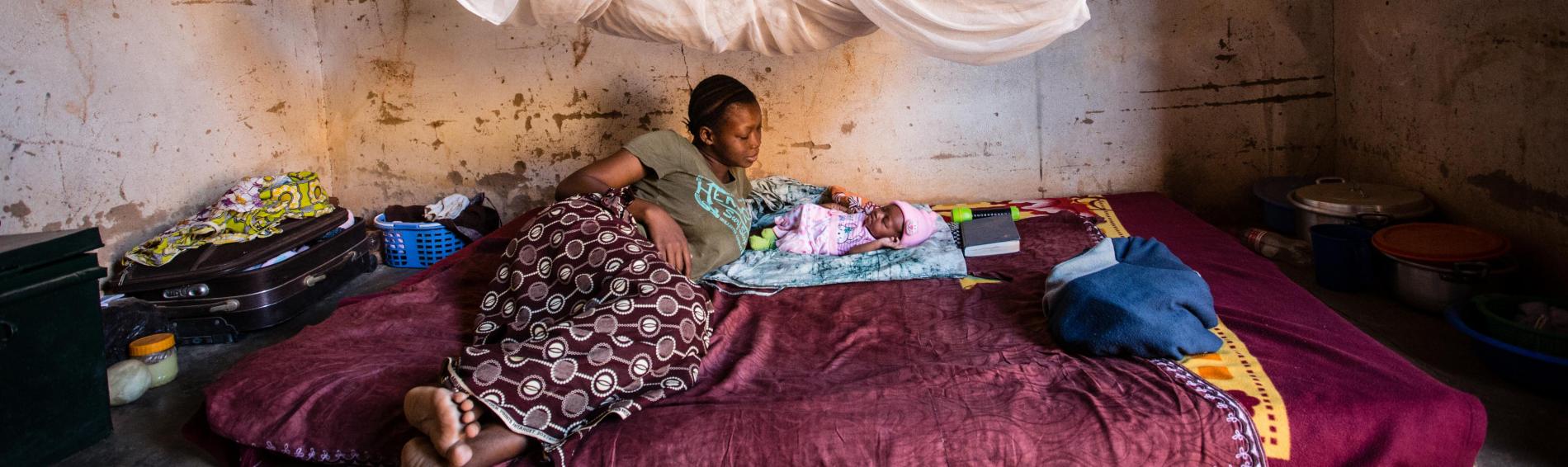 Een Burkinese tienermoeder in kleurrijke jurk ligt uitgestrekt naast haar zuigeling op een matras tussen een koffer, potten en pannen. De kamer is erg kaal. Aan het plafond hangt een muskietennet.