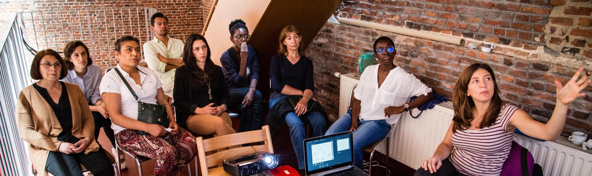 Een groep vrouwen van verschillende leeftijd en achtergrond zit op stoelen en kijkt naar een projectie. Op een stoel staat een laptop. 