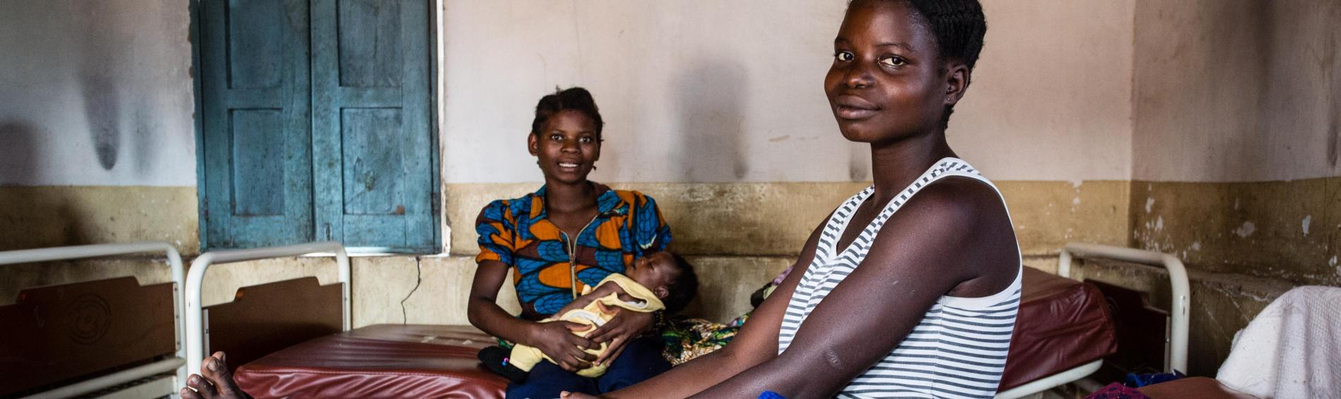 Twee Congolese vrouwen in kleurrijke jurken zitten op ziekenhuisbedden, hun zuigelingen in de arm.