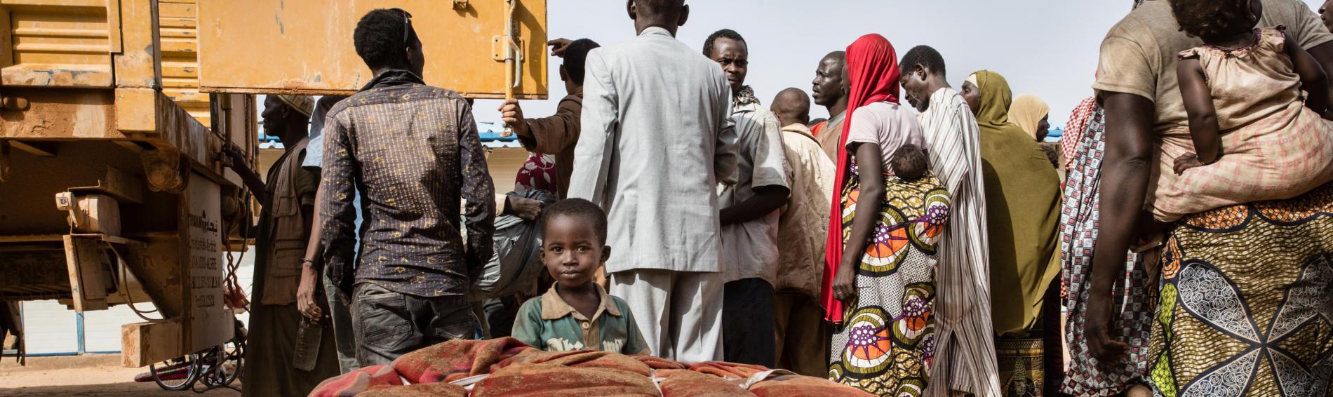 Een groep Nigeriaanse mannen, vrouwen en kinderen staat rond een vrachtwagen die wordt uitgeladen. Op de voorgrond kijkt een jongetje in de lens, vanachter een zware vracht die naast de menigte is neergeploft. 
