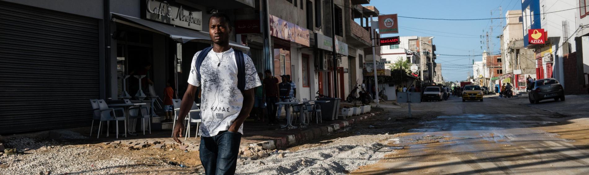 Een jongeman van Afrikaanse orgine loopt door een verlaten straat in Tunis. Hij draagtt rode sportschoenen, een jeans en een T-shirt waarop 'Ellada' (Griekenland) staat geschreven. Op de achtergrond zijn winkels te zien. De straat is in slechte staat en ligt er verlaten bij.