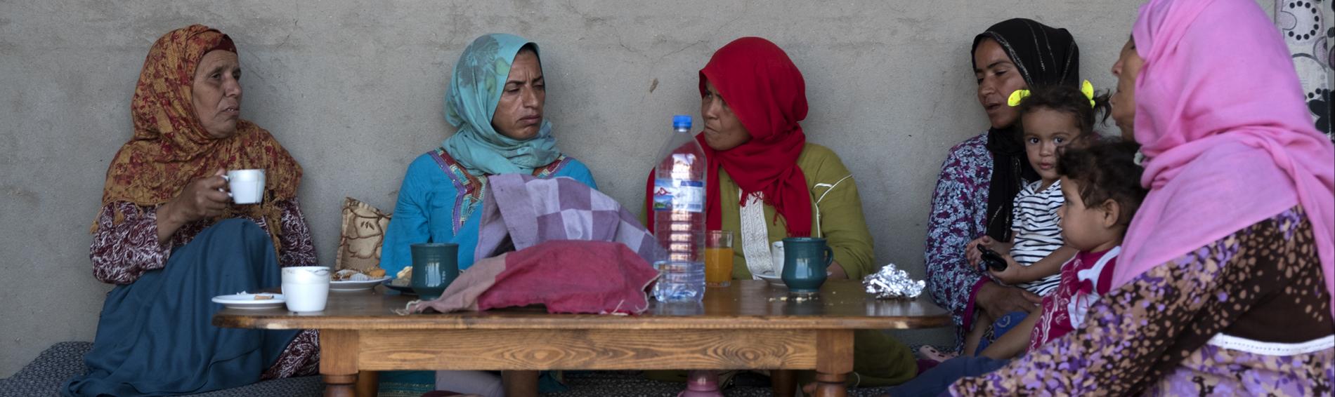 Vijf vrouwen van uiteenlopende leeftijden met kleurrijke hiqabs en jurken, een meisje met twee staartjes en een jongetje, zitten tegen een muur buiten een huis in Tunesië. Ze zitten op kussens rond een lage tafel waarop water, thee en koffie staat. 