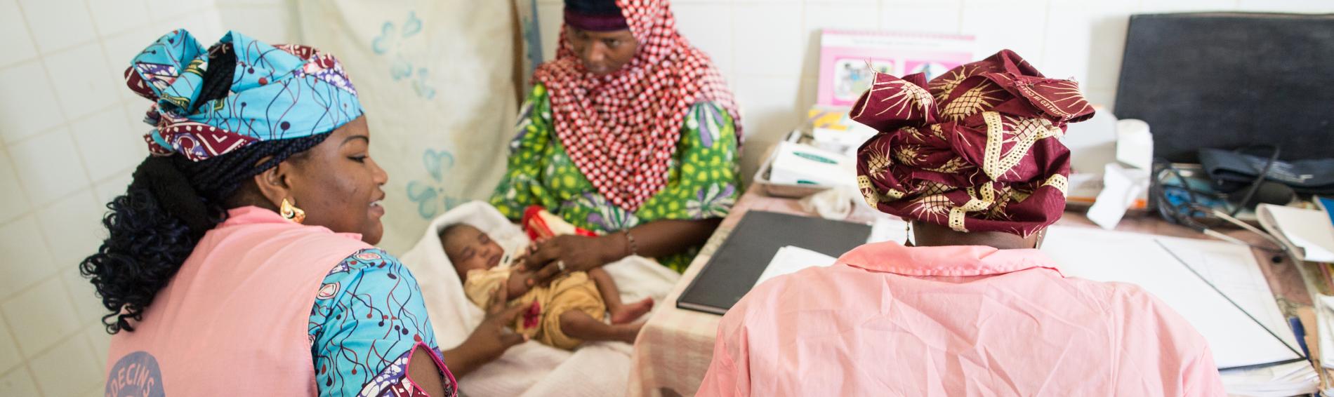 Twee Nigeriaanse medewerksters van Dokters van de Wereld met roze vesten waarop het logo staat, behandelen een vrouw en haar baby in ons centrum in Agadez. 