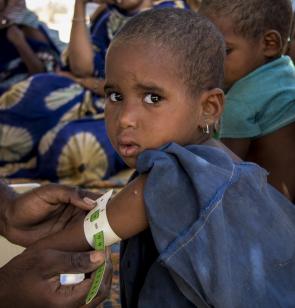Een Malinees meisje kijkt recht in de camera terwijl een arts de omtrek van haar verzwakte armpje meet.